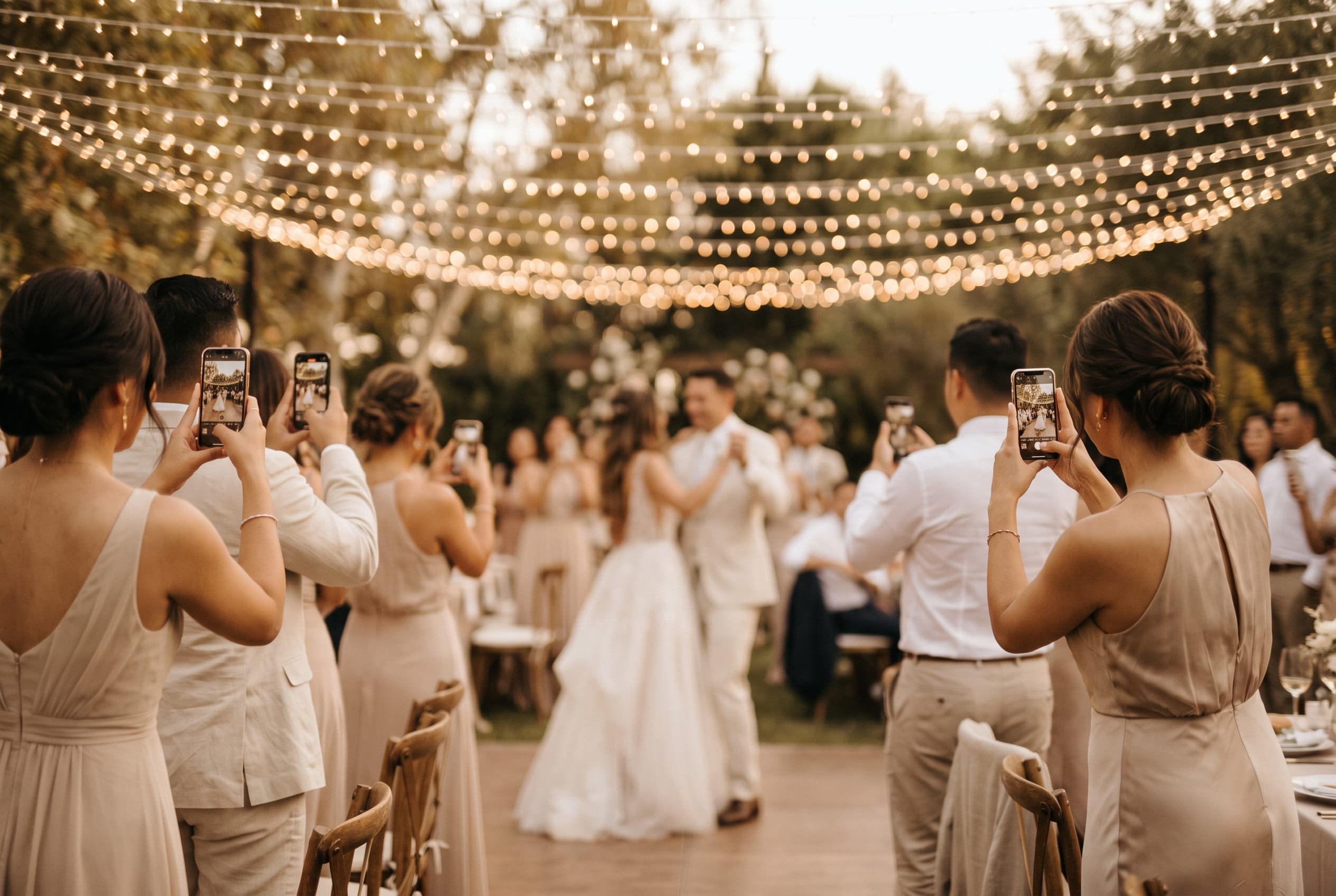 Invités de mariage photographiant les mariés qui dansent au crépuscule, guirlande lumineuse au-dessus de la piste