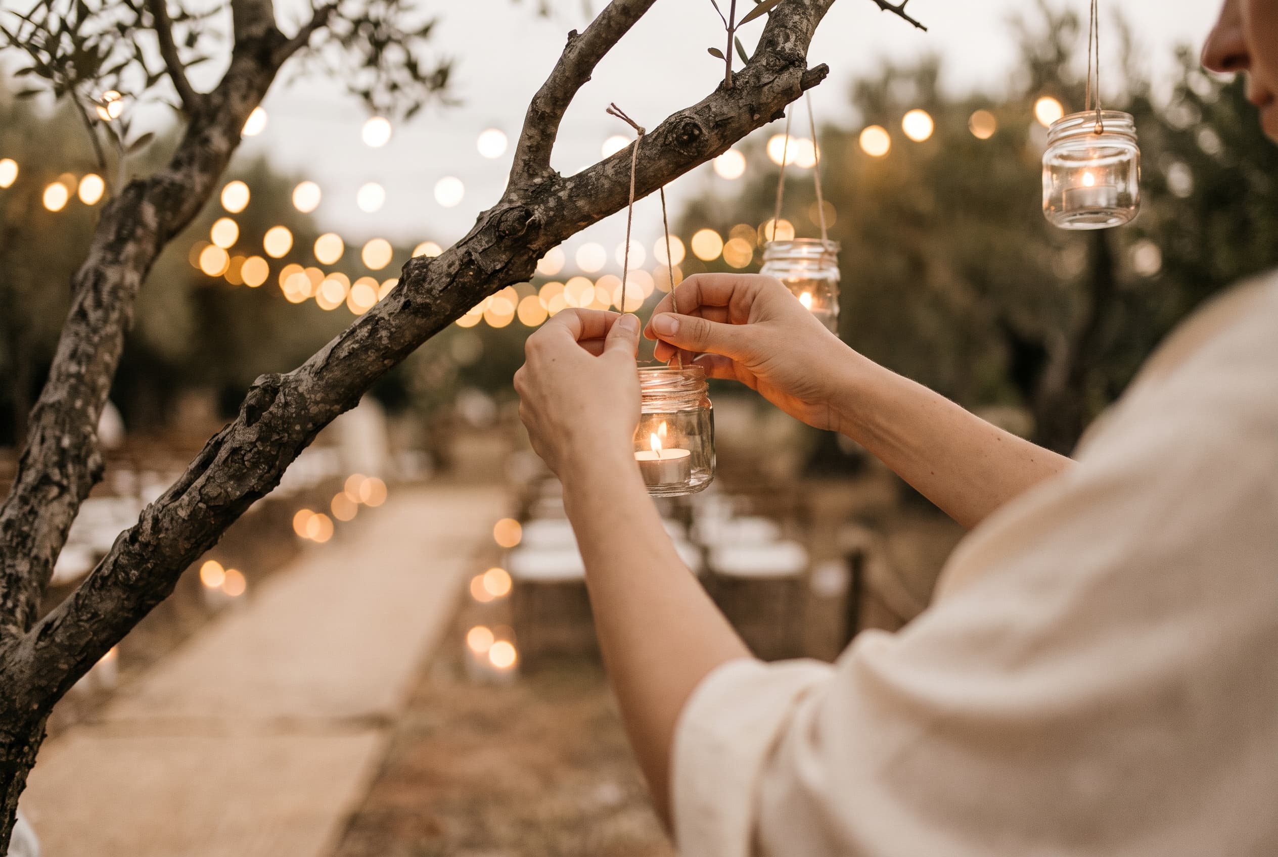 Bocaux en verre recyclés suspendus à une branche d'arbre avec une bougie à l'intérieur, éclairant une allée de cérémonie de mariage au crépuscule