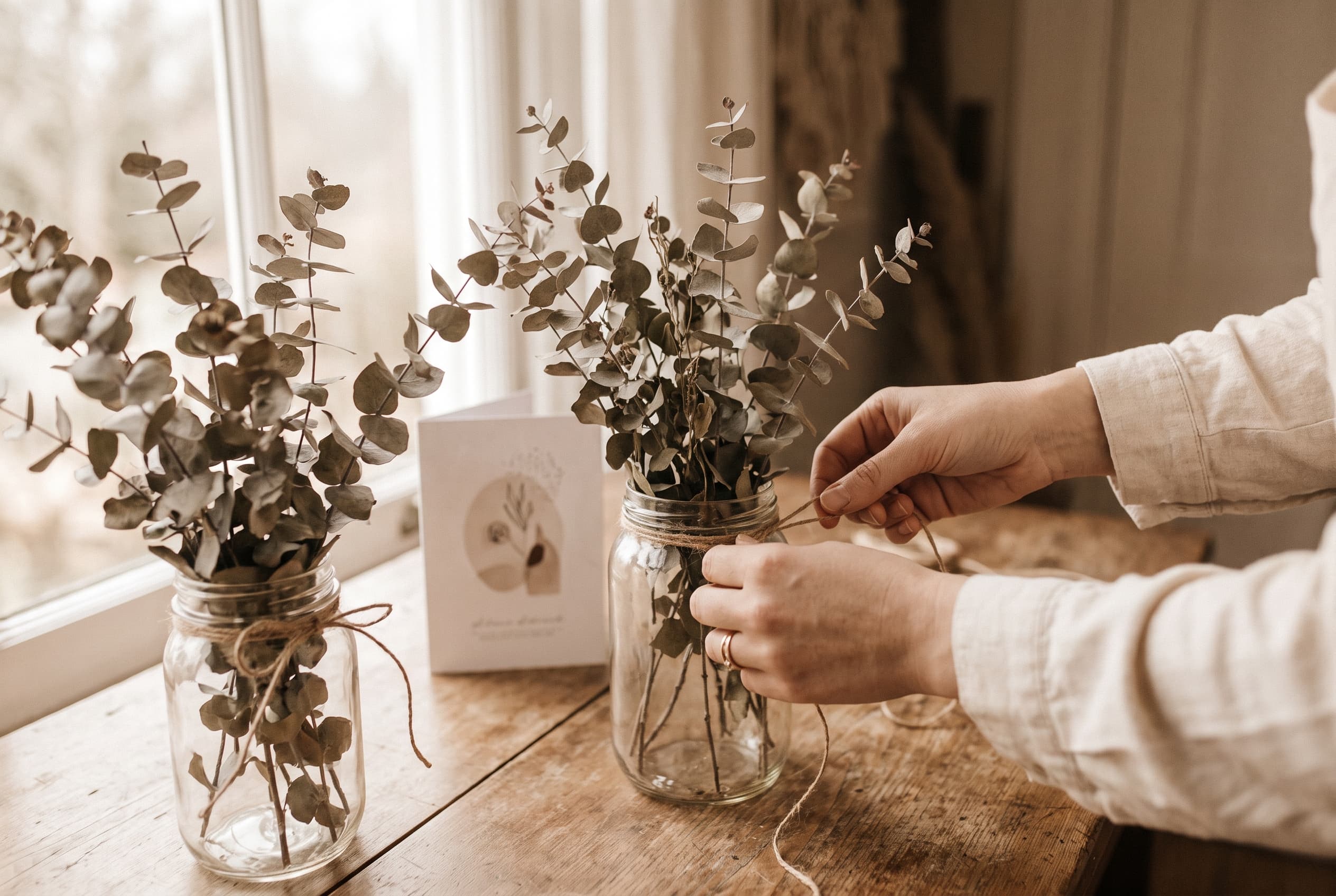 Mains assemblant un centre de table de mariage à partir de bocaux en verre récupérés et de branches d'eucalyptus séché sur une table en bois brut