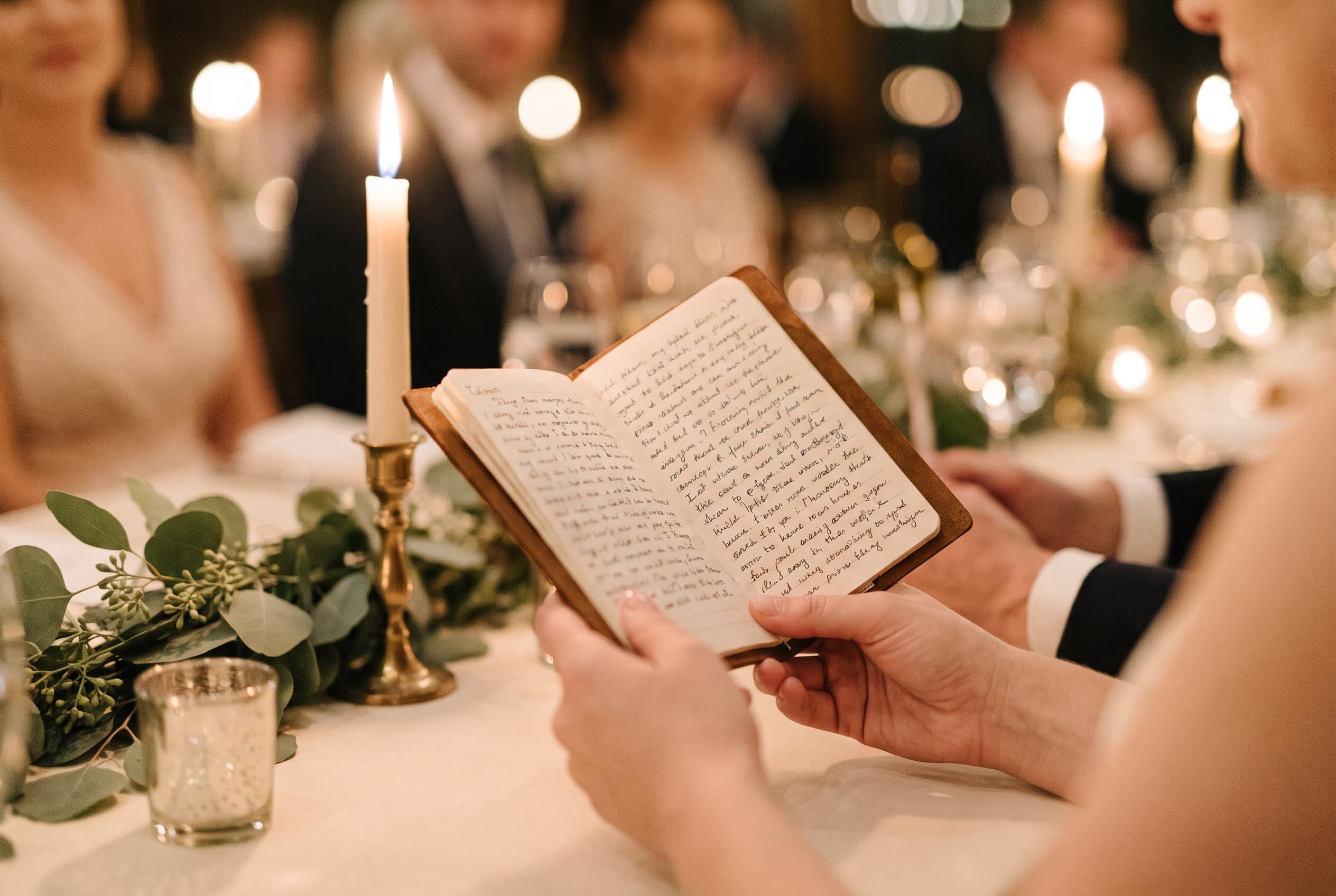 Gros plan sur des mains tenant un carnet avec des notes manuscrites de discours, posé sur une table de réception éclairée à la bougie