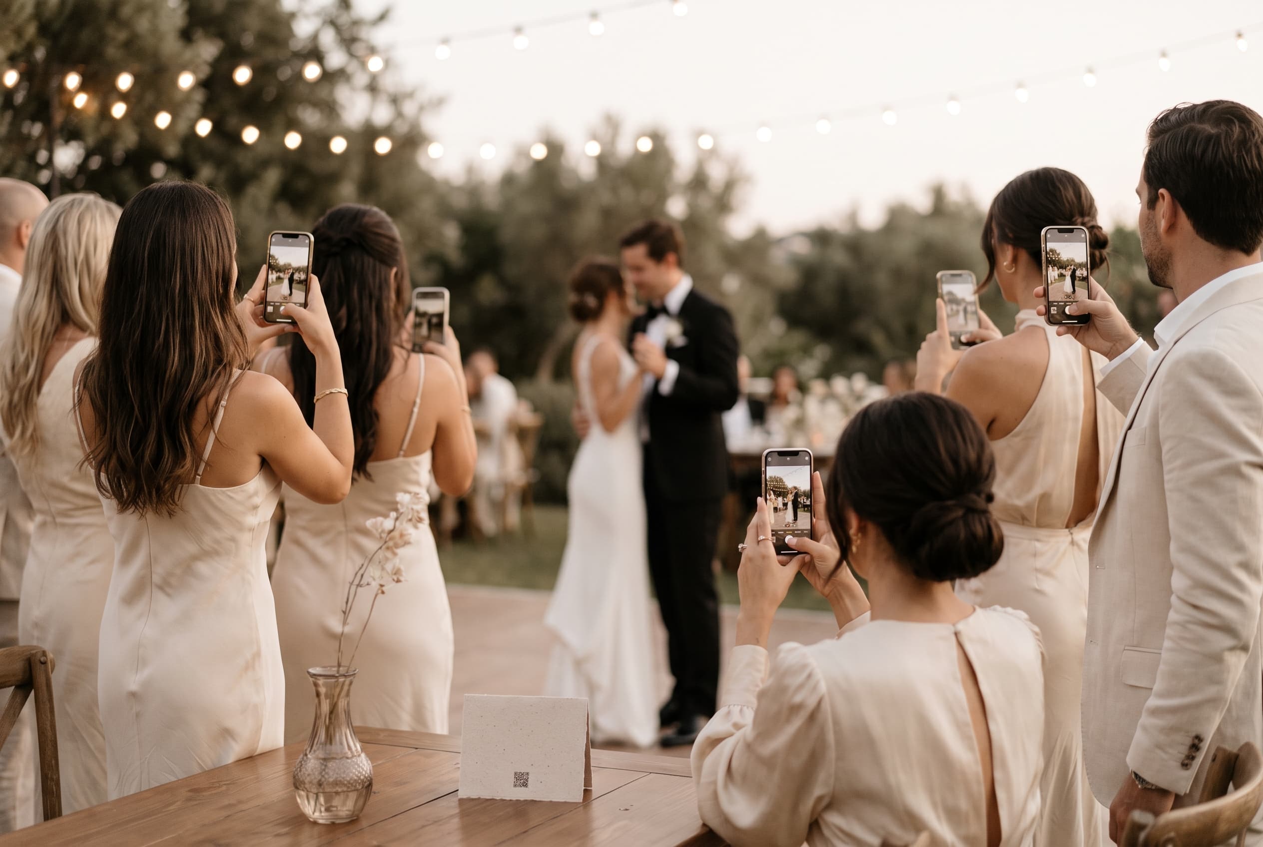 Invités de mariage photographiant les mariés qui dansent au crépuscule, guirlande lumineuse au-dessus de la piste