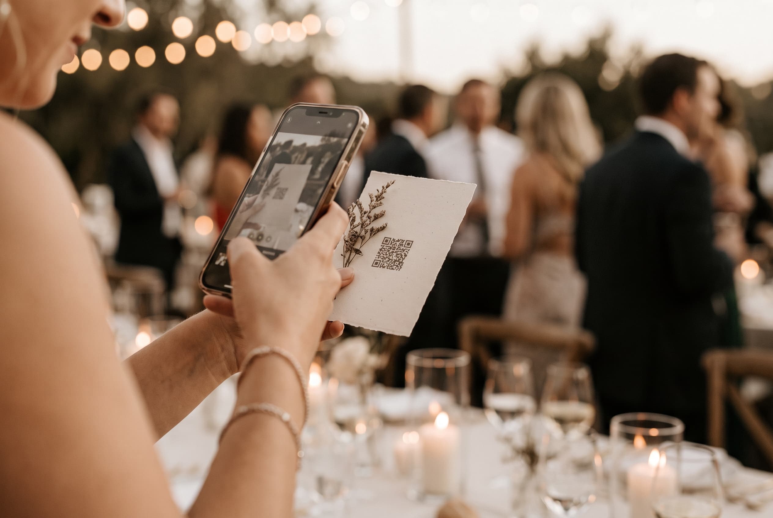 Invitée de mariage photographiant un QR code posé sur une table décorée de bougies lors d'un cocktail en extérieur