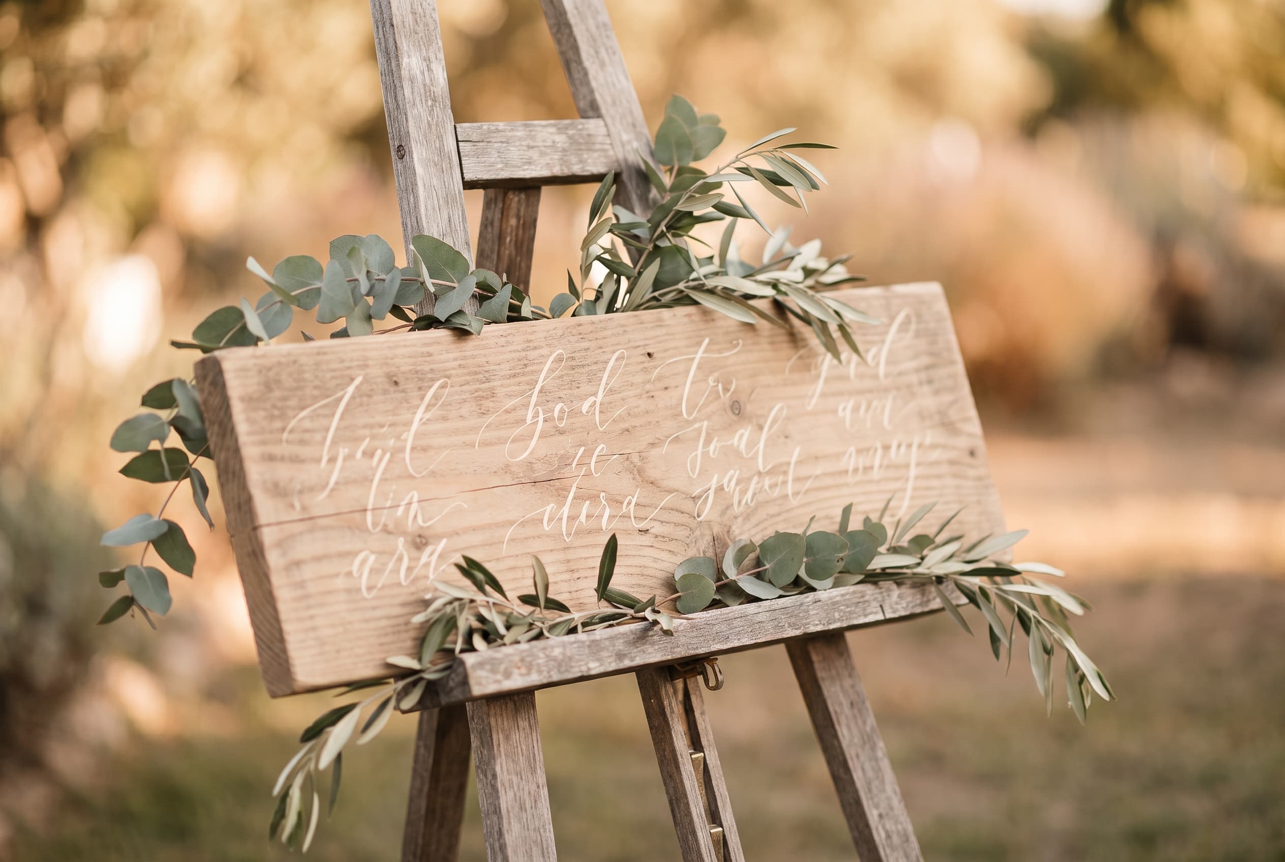 Plan de table de mariage sur une planche de palette poncée, posée sur un chevalet en bois, calligraphie à la craie, brins d'eucalyptus, lumière dorée