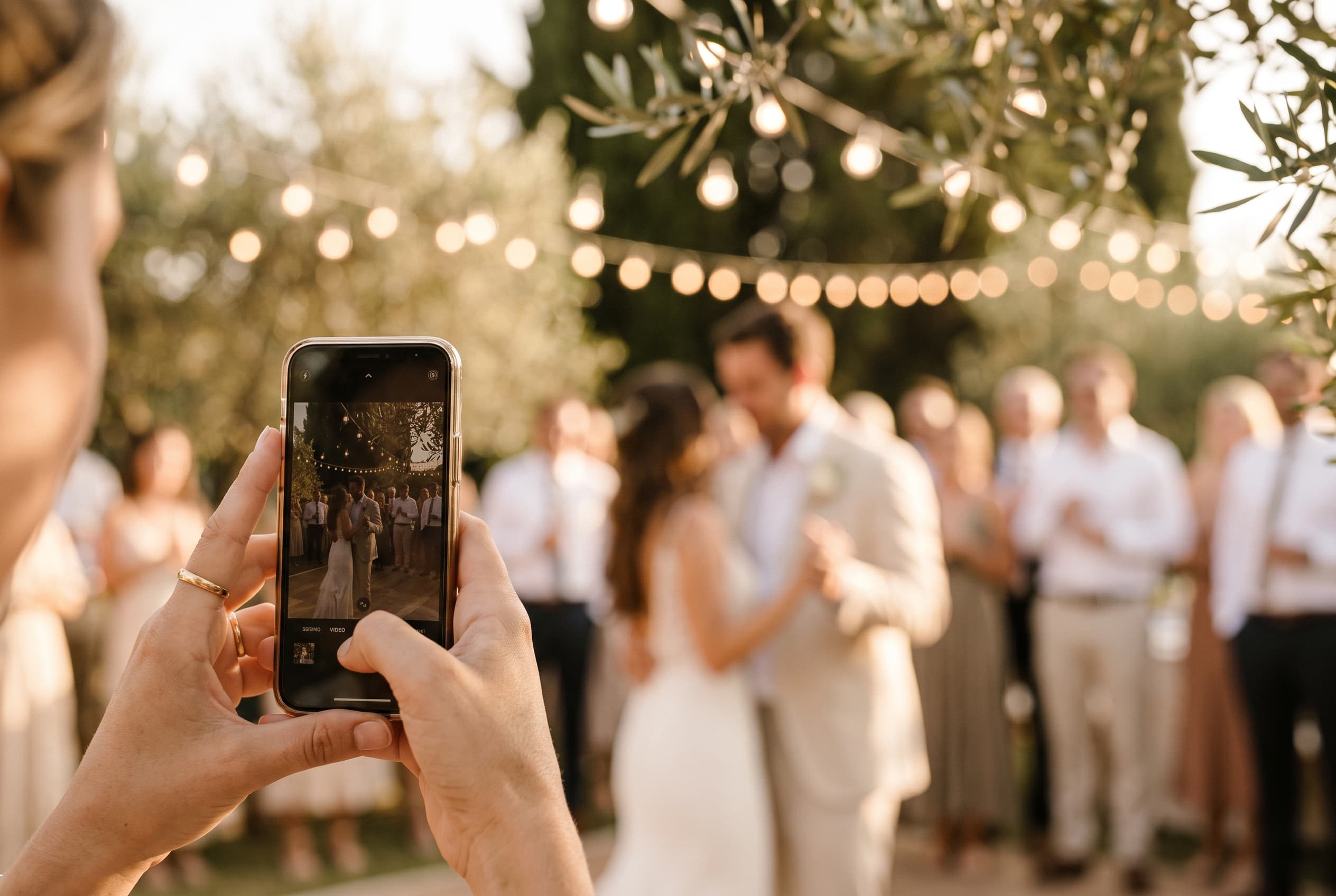 Main d'un invité de mariage tenant un smartphone lors d'une réception en extérieur à la lumière dorée du soir